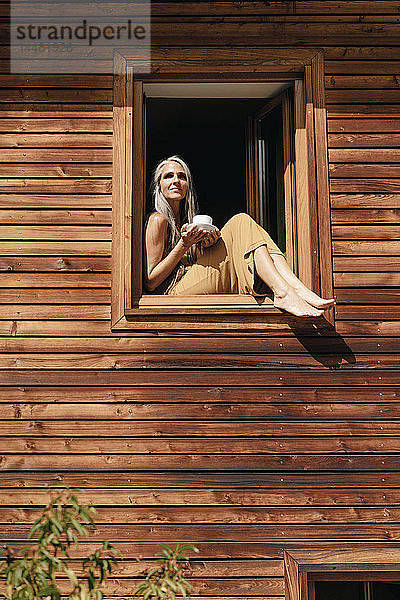 Frau mit Tasse Kaffee sitzt auf dem Fensterbrett ihres Hauses und sonnt sich