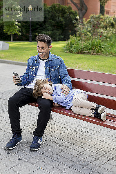 Glücklicher Vater mit Sohn auf einer Bank mit Handy und Ohrstöpseln