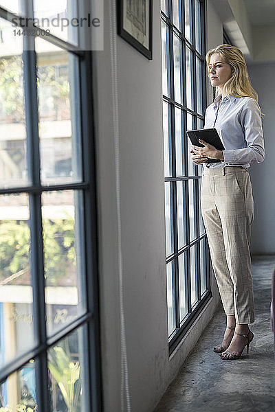 Selbstbewusste Geschäftsfrau mit Tablett  die im Büro steht und aus dem Fenster schaut