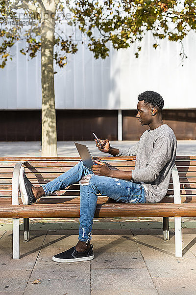 Junger Mann sitzt auf einer Bank in der Stadt und benutzt Laptop und Smartphone