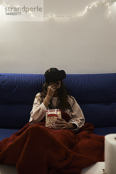 Junge Frau mit VR-Headsets isst nachts zu Hause Popcorn auf dem Sofa