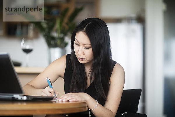 Junge Frau in schwarzem Kleid benutzt Laptop und macht Notizen auf dem Tisch zu Hause