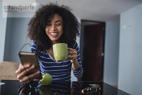 Porträt einer glücklichen jungen Frau mit Kaffeetasse beim Blick aufs Handy