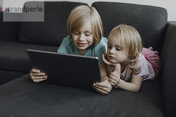 Portrait von Bruder und kleiner Schwester  die zu Hause auf der Couch liegen und ein digitales Tablet benutzen