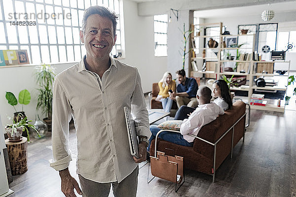Porträt eines lächelnden reifen Geschäftsmannes mit Mitarbeitern im Hintergrund im Loft-Büro