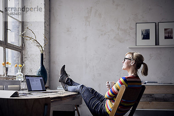 Lächelnde Frau sitzt mit erhobenen Füßen am Schreibtisch auf einem Dachboden und schaut durchs Fenster