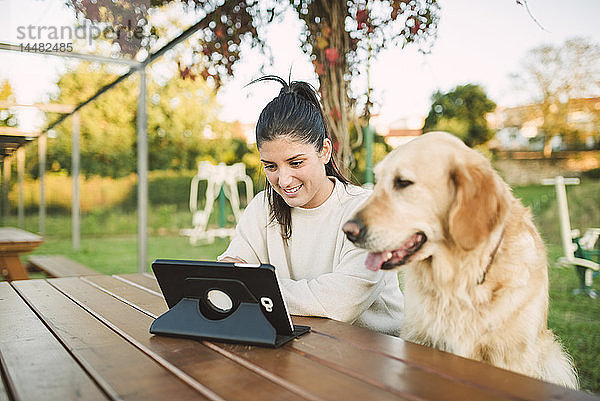 Lächelnde junge Frau mit einer Tablette in einem Park mit ihrem Hund