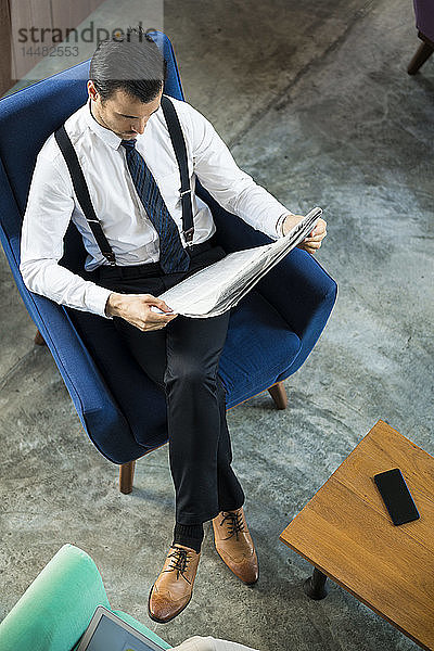 Draufsicht auf einen Geschäftsmann  der in einem blauen Sessel sitzt und Zeitung liest