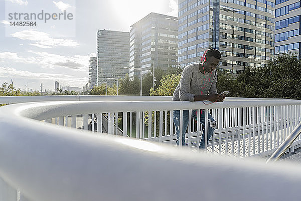 Junger Mann steht auf einer Brücke  hört Musik  benutzt ein Smartphone