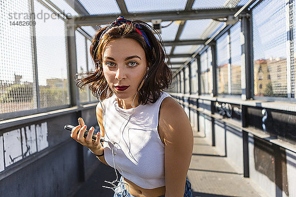 Porträt einer jungen Frau mit Ohrstöpseln und Handy auf einer Brücke