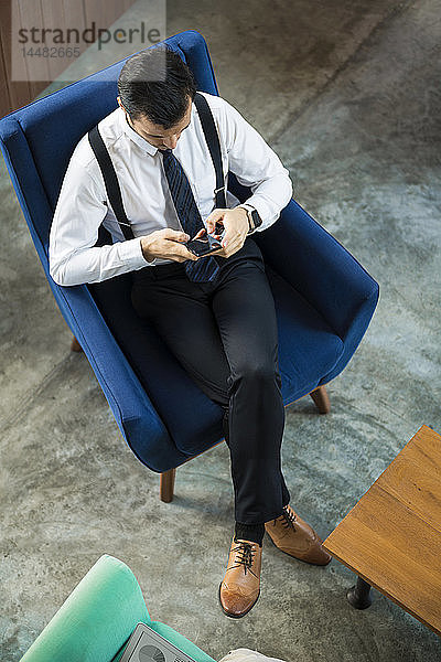 Draufsicht eines Geschäftsmannes  der in einem blauen Sessel sitzt und ein Smartphone benutzt