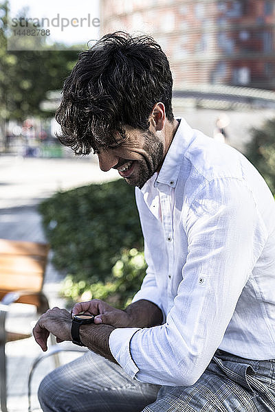 Lächelnder junger Mann sitzt mit Smartwatch im Freien