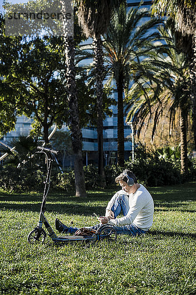 zufälliger Geschäftsmann  der auf einer Wiese in einem Park sitzt und ein Smartphone und Kopfhörer benutzt