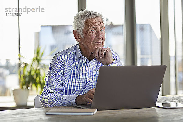Porträt eines nachdenklichen älteren Mannes  der mit Laptop am Tisch sitzt und in die Ferne schaut