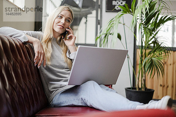 Lächelnde junge Frau sitzt auf der Couch mit Laptop und telefoniert mit dem Handy