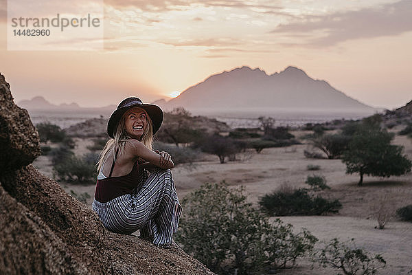 Namibia  Spitzkoppe  lachende Frau  die bei Sonnenuntergang auf einem Felsen sitzt