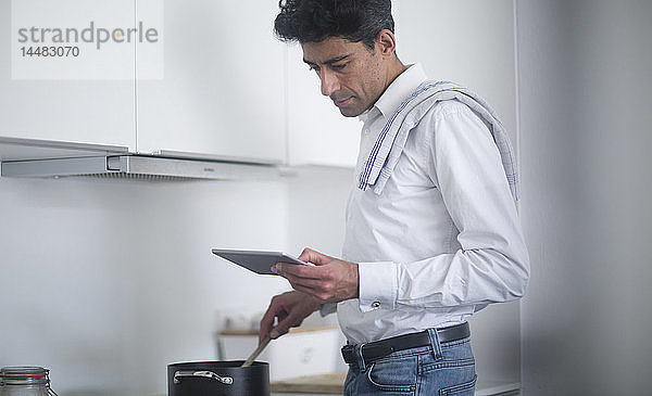Mann steht in der Küche und liest während des Kochens Nachrichten auf seinem digitalen Tablet