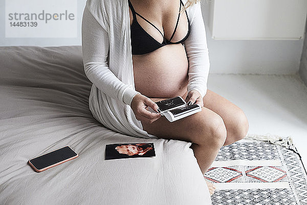 Schwangere Frau zu Hause am Bett sitzend mit Ultraschallbildern