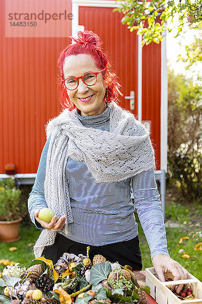 Porträt einer lächelnden älteren Frau mit rot gefärbten Haaren beim Basteln an der Herbstdekoration im Garten