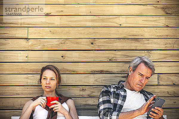 Frau mit Kaffeetasse und Mann mit Smartphone vor einer Blockhütte