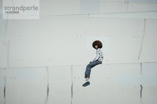 Porträt selbstbewusster junger Mann mit Afro  der auf einer sonnigen Stadtmauer sitzt