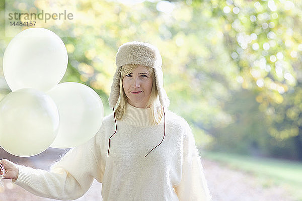 Porträt selbstbewusste Frau mit weißer Pelzmütze  die ein Bündel Luftballons im Park hält