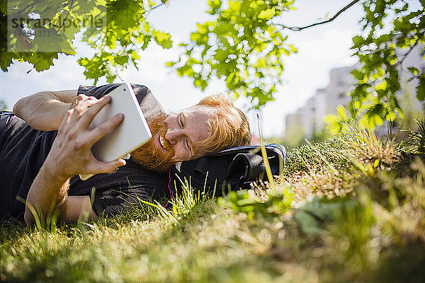 Lächelnder Mann mit Bart  der in einem sonnigen Park ein digitales Tablet benutzt