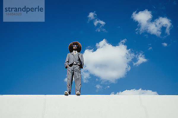 Selbstbewusster  cooler  gut gekleideter junger Mann mit Afro  der an einer sonnigen Wand vor blauem Himmel steht