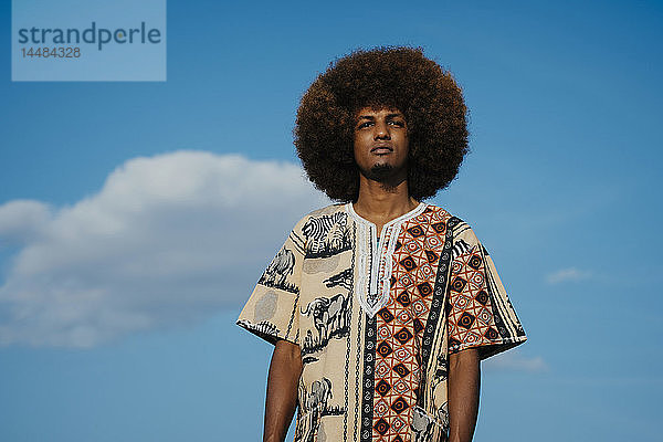 Porträt selbstbewusster junger Mann mit Afro gegen blauen Himmel mit Wolken stehend