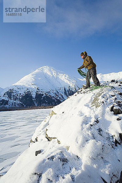 Ein Bergsteiger steht in der Nähe der Spitze einer Klippe über der gefrorenen Oberfläche des Portage Lake im Chugach National Forest. Winter in Süd-Zentral-Alaska.