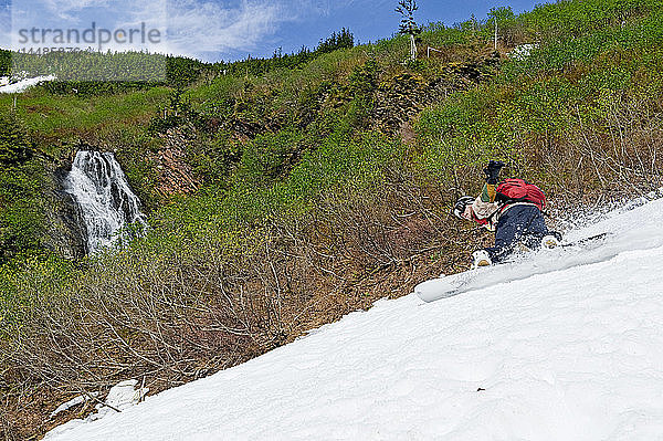 Ein Snowboarder fährt eine Kurve im Frühlingsschnee mit einem von der Schneeschmelze gespeisten Wasserfall im Hintergrund in der Nähe von Juneau  Alaska