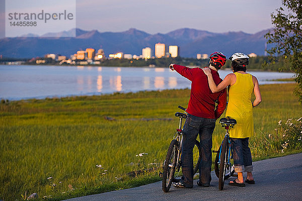 Radfahrer  die sich ausruhen und den Sonnenuntergang entlang des Tony Knowles Coastal Trail mit der Skyline von Anchorage im Hintergrund genießen  Southcentral Alaska