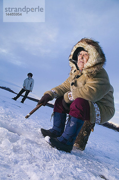 Ältere einheimische Yupik-Frau sitzt auf einem Baumstumpf Eisfischen Kuskokwim River Tuluksak Westalaska Winter