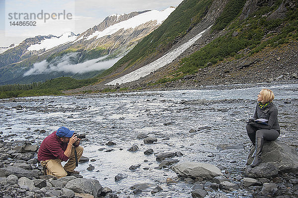 Mann fotografiert eine Frau  die auf einem Felsen im Portage Creek  Portage Valley  Süd-Zentral-Alaska  sitzt und in ihr Tagebuch schreibt