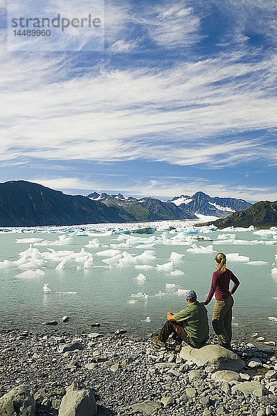 Ein Paar  das im Kenai Fjords National Park wandert  ruht sich aus und betrachtet den Bear Glacier vom Ufer des Bear Glacier Lake  Kenai Peninsula  Southcentral Alaska  Sommer