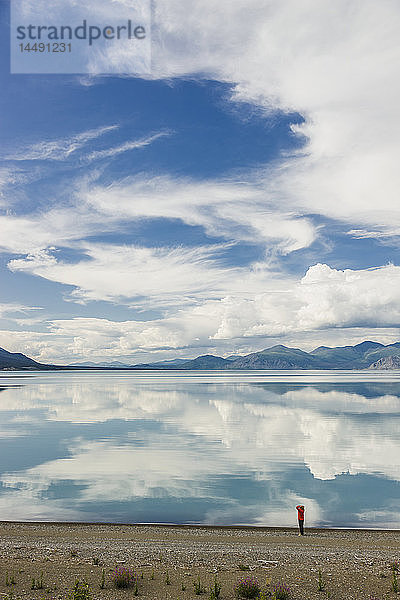 Eine Frau steht am Ufer des Kluane Lake mit Bergen und Wolken  die sich im ruhigen Wasser des Kluane Lake spiegeln  Yukon Territorium  Kanada  Sommer