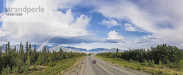 Ein Auto fährt auf dem Alaska Highway neben dem Kluane Lake mit atmosphärischen Regenwolken und einem Regenbogen im Hintergrund  Yukon Territory  Kanada  Sommer