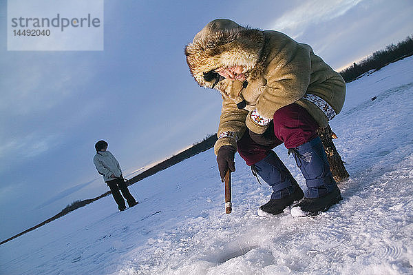 Ältere einheimische Yupik-Frau sitzt auf einem Baumstumpf Eisfischen Kuskokwim River Tuluksak Westalaska Winter