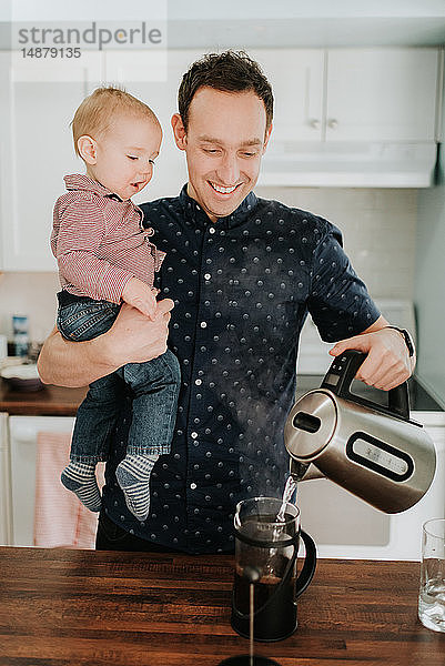 Vater trägt Baby Sohn Sohn macht Kaffee an der Küchentheke