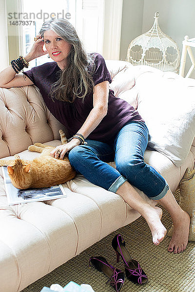 Stilvolle reife Frau sitzt auf dem Sofa und streichelt Ingwer-Katze  Porträt