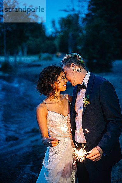 Romantische Braut und Bräutigam mit Wunderkerzen am Seeufer in der Abenddämmerung