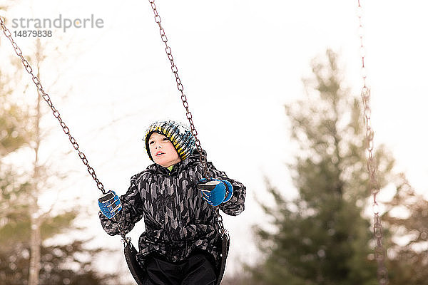 Junge mit Strickmütze schwingt im Winter auf der Spielplatzschaukel