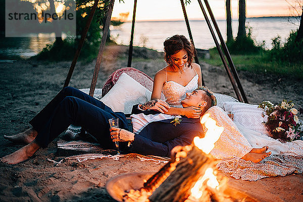 Braut und Bräutigam liegend am Lagerfeuer am Seeufer in der Abenddämmerung