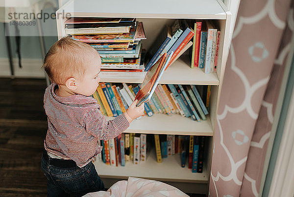 Kleiner Junge im Wohnzimmer nimmt Geschichtenbuch aus Bücherregalen