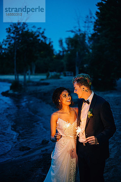 Romantisches Brautpaar mit Wunderkerzen beim Spaziergang am Seeufer in der Abenddämmerung