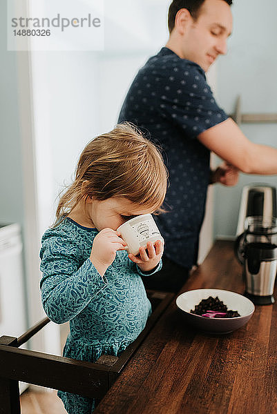 Weibliches Kleinkind mit Vater trinkt aus Tasse an der Küchentheke