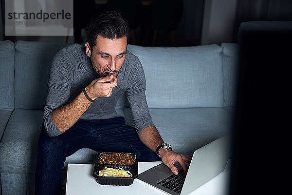 Junger Mann sitzt abends auf dem Sofa  isst zum Mitnehmen und benutzt Laptop