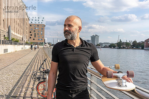 Mann hält Skateboard auf Brücke  Fluss und Gebäuden im Hintergrund  Berlin  Deutschland