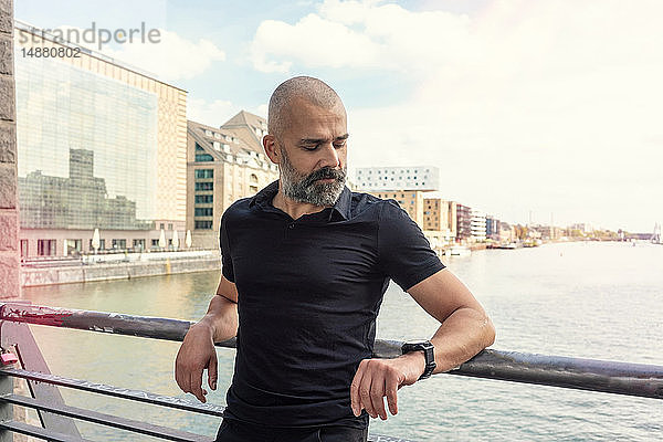 Mann betrachtet Smartwatch auf Brücke  Fluss und Gebäude im Hintergrund  Berlin  Deutschland