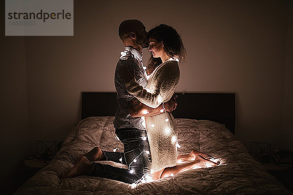 Romantisches junges Paar in dekorativen Lichtern verschlungen kniend von Angesicht zu Angesicht auf dem Bett